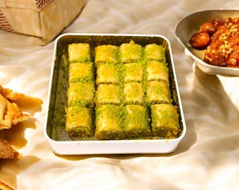 May include: A rectangular white tin filled with fifteen square pieces of golden-brown baklava, each topped with a generous dusting of bright green crushed pistachios. The pastry is arranged in a neat grid pattern within the container.