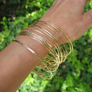 May include: A stack of gold-toned bangle bracelets on a person's wrist. The bracelets are thin and have a hammered texture.