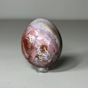 May include: A polished, egg-shaped stone with a mix of colors, including burgundy, white, and tan. The stone sits on a clear, round stand. The surface has a marbled appearance, with veins of color running throughout.