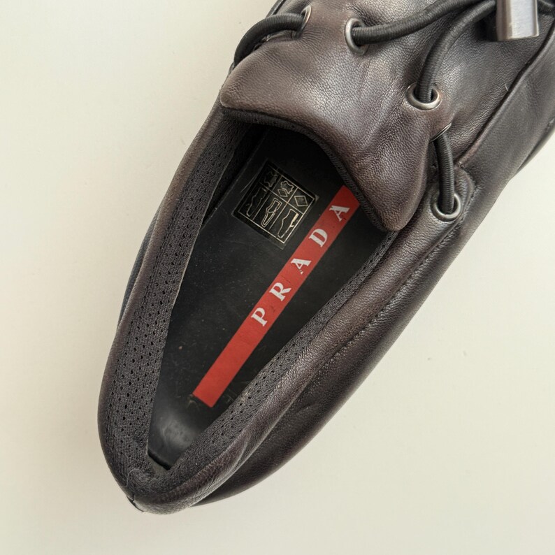 Puede incluir: Primer plano de un mocas&iacute;n de cuero marr&oacute;n con cordones negros. El interior presenta una franja roja con el nombre de la marca "PRADA" en letras blancas. El zapato est&aacute; dise&ntilde;ado con una puntera redondeada y un perfil bajo.