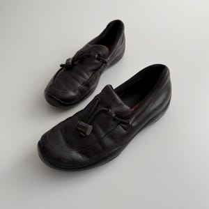 Puede incluir: Un par de mocasines de cuero negro con punta redondeada y detalle de lazo decorativo. Los zapatos tienen un tac&oacute;n bajo y una plantilla acolchada. Los mocasines son un estilo cl&aacute;sico, adecuado para uso informal o semiformal.