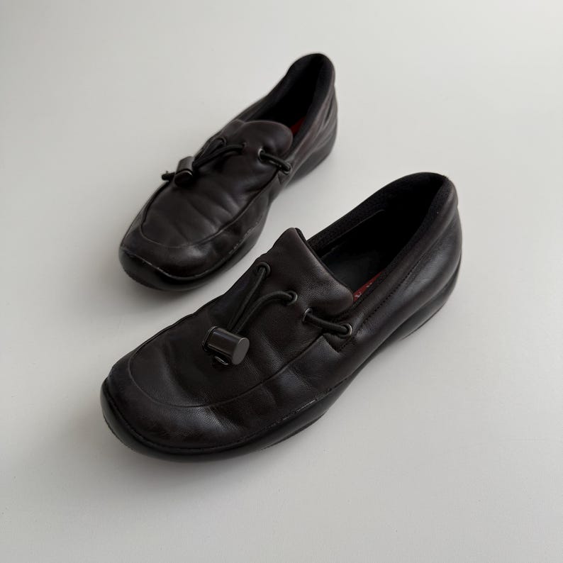 Puede incluir: Un par de mocasines de cuero negro con punta redondeada y detalle de lazo decorativo. Los zapatos tienen un tac&oacute;n bajo y una plantilla acolchada. Los mocasines son un estilo cl&aacute;sico, adecuado para uso informal o semiformal.