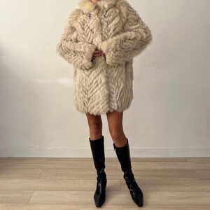 Pu&ograve; includere: Un cappotto in finta pelliccia color crema con un motivo a spina di pesce. Il cappotto ha un colletto e maniche lunghe. La persona indossa stivali neri al ginocchio.