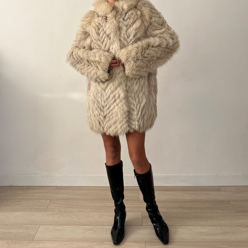 Pu&ograve; includere: Un cappotto in finta pelliccia color crema con un motivo a spina di pesce. Il cappotto ha un colletto e maniche lunghe. La persona indossa stivali neri al ginocchio.