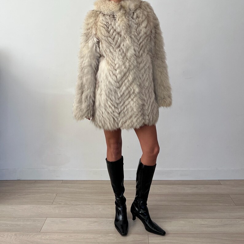 Pu&ograve; includere: Un cappotto di pelliccia colorata, lungo fino al ginocchio, con un motivo a spina di pesce. Il cappotto ha un ampio colletto e maniche lunghe. La persona indossa stivali neri al ginocchio con la punta.