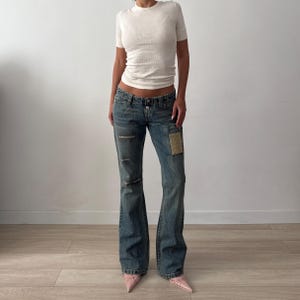 Op de afbeelding: Een wit T-shirt met korte mouwen en ronde hals gecombineerd met een blauwe spijkerbroek met lage taille. De broek heeft een versleten afwerking met horizontale scheuren op het rechterbeen en een rechthoekige beige patch op de linkerdij, met knoopsluiting.