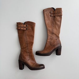 Puede incluir: Un par de botas altas de cuero color canela con tacón de bloque, punta redondeada y detalles perforados decorativos a lo largo de la caña. Cada bota incluye dos correas ajustables con hebillas metálicas en la parte superior y un degradado.