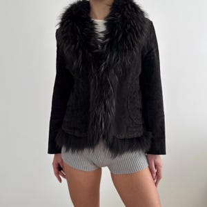 May include: A black suede jacket with a faux fur collar and trim. The jacket has long sleeves and two front pockets. The faux fur is black and grey. The jacket is a classic style.
