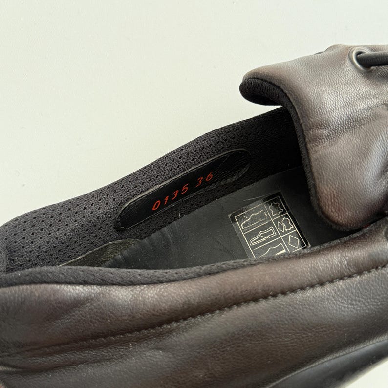 Puede incluir: Primer plano de un zapato de cuero marr&oacute;n. El interior del zapato revela un forro de malla negra y una etiqueta con los n&uacute;meros "0135 36". El zapato tiene un exterior marr&oacute;n oscuro y una suela negra.