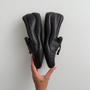 Puede incluir: Un par de mocasines de cuero negro. Los zapatos tienen una punta redondeada, un tac&oacute;n bajo y un detalle decorativo de lazo en la pala. Los zapatos est&aacute;n sostenidos por una mano.