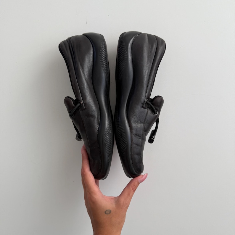 Puede incluir: Un par de mocasines de cuero negro. Los zapatos tienen una punta redondeada, un tac&oacute;n bajo y un detalle decorativo de lazo en la pala. Los zapatos est&aacute;n sostenidos por una mano.