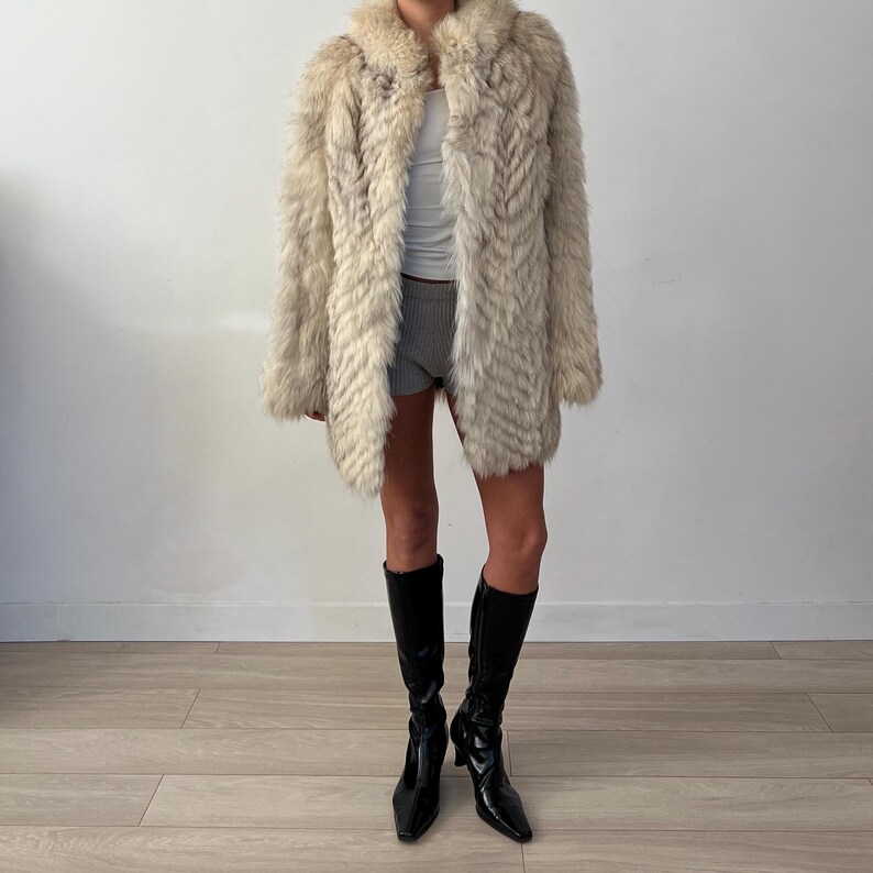 Pu&ograve; includere: Un cappotto di pelliccia color crema con un motivo a spina di pesce. Il cappotto ha un ampio colletto e maniche lunghe. La persona indossa stivali neri al ginocchio.