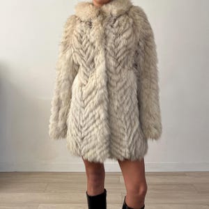 Pu&ograve; includere: Un cappotto di pelliccia colorata con un motivo a spina di pesce. Il cappotto ha un colletto e maniche lunghe. La pelliccia appare morbida e soffice, con variazioni di colore. Il cappotto &egrave; lungo fino al ginocchio.