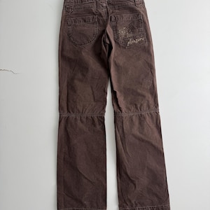 May include: Dark brown pants featuring a horizontal seam across the knees, belt loops, and two back pockets. The right back pocket displays a stitched graphic of a butterfly and the word pleasure in cursive script, with visible topstitching.