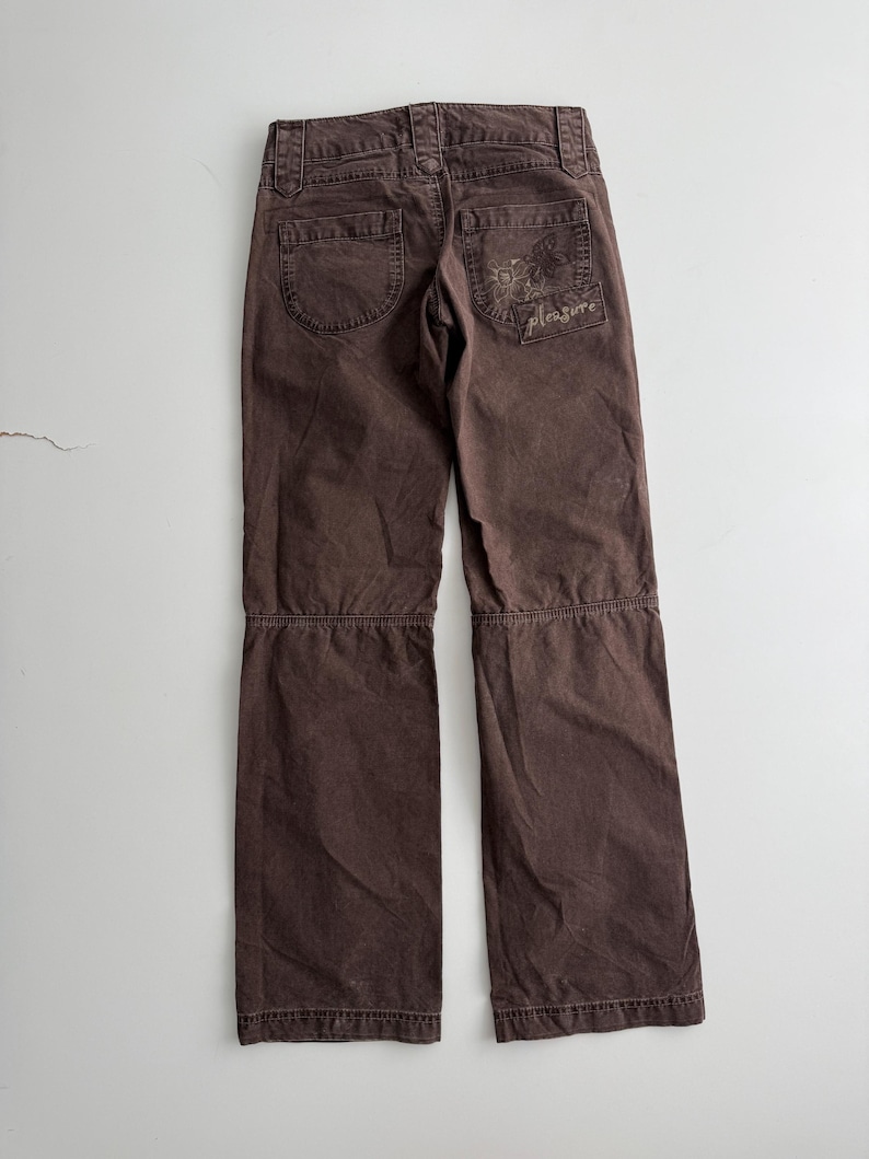 May include: Dark brown pants featuring a horizontal seam across the knees, belt loops, and two back pockets. The right back pocket displays a stitched graphic of a butterfly and the word pleasure in cursive script, with visible topstitching.