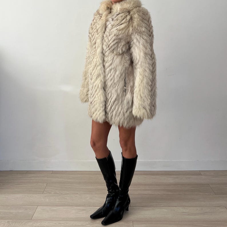 Pu&ograve; includere: Un cappotto di pelliccia vintage, lungo fino al ginocchio, nei toni del crema e del grigio. Il cappotto presenta un motivo strutturato a spina di pesce e un ampio colletto. La modella indossa stivali neri al ginocchio.