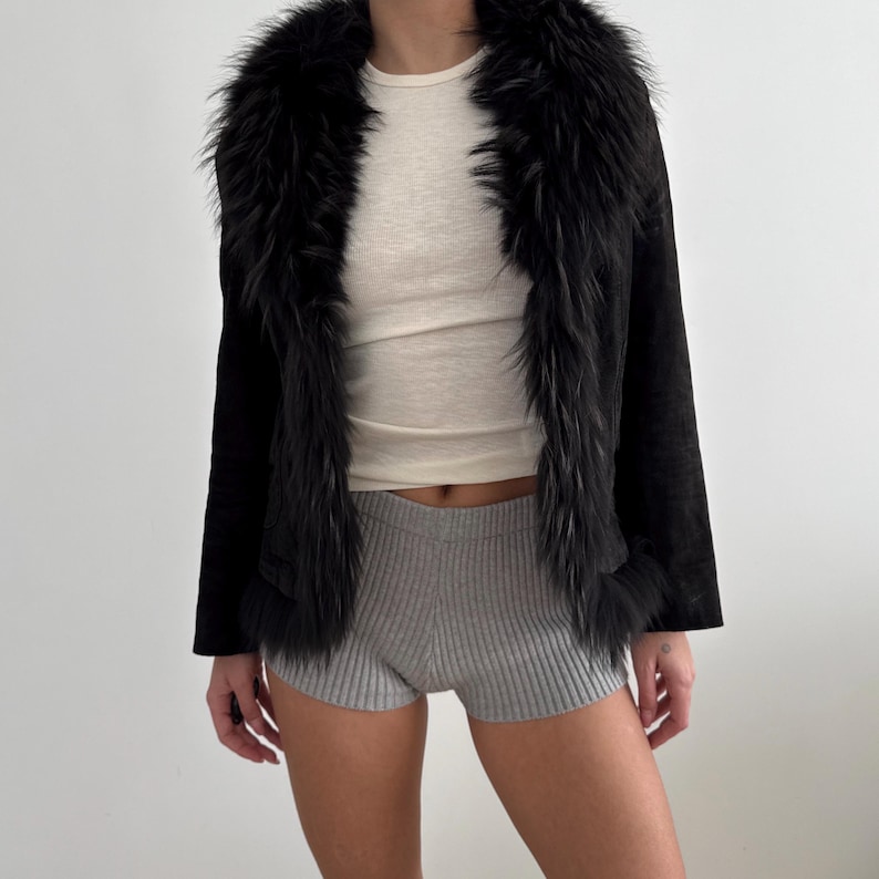 May include: A black jacket featuring a faux fur collar and trim. The jacket is paired with a cream-coloured top and grey shorts. The fur trim on the jacket adds a touch of luxury to the outfit.