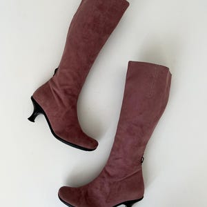 May include: A pair of knee-high boots in a dusty rose color. The boots have a soft, suede-like texture and feature a small, black heel. A zipper is visible on the back of each boot. The boots are displayed on a white background.