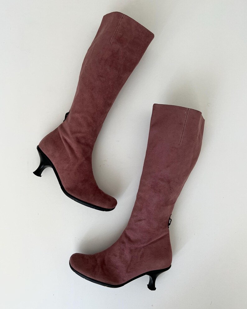 May include: A pair of knee-high boots in a dusty rose color. The boots have a soft, suede-like texture and feature a small, black heel. A zipper is visible on the back of each boot. The boots are displayed on a white background.