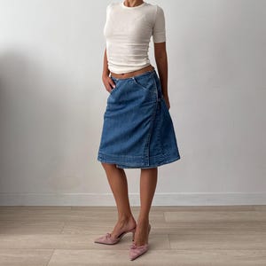 May include: A white short-sleeved crew neck top paired with a knee-length blue denim skirt featuring a wrap-style front, visible stitching, and a low-rise waistline. The outfit is completed with a pair of light pink pointed-toe heeled mules.