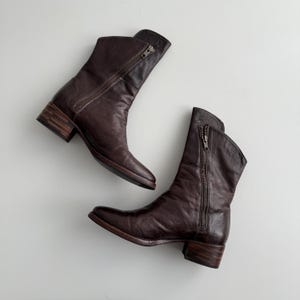 May include: Two dark brown leather ankle boots with side zips. The boots feature a low, stacked heel and a pointed toe. The leather has a worn, textured appearance with creases. The boots are displayed on a white surface.