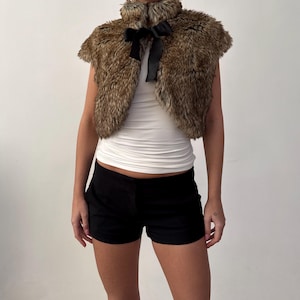 May include: A brown faux fur bolero with a black satin bow. The short-sleeved jacket has a high collar and is worn over a white tank top. Black shorts complete the outfit.