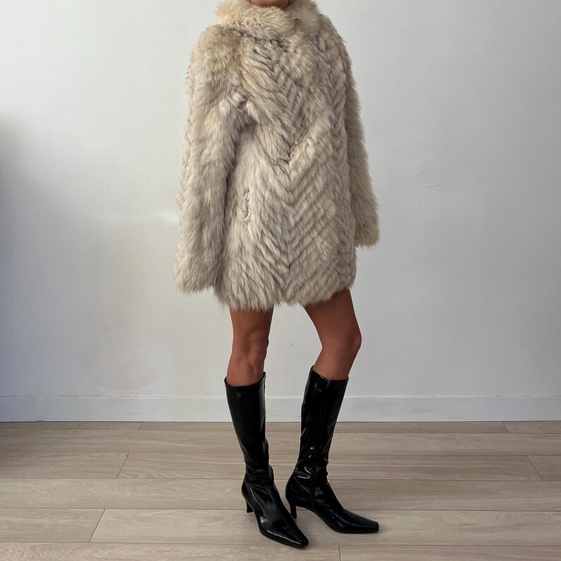 Pu&ograve; includere: Un cappotto di pelliccia chiara e strutturata con colletto. Il cappotto ha un motivo a spina di pesce e viene indossato con stivali neri al ginocchio con punta e tacchi piccoli.