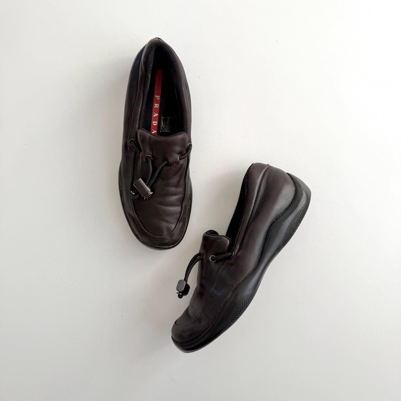 Puede incluir: Dos mocasines de cuero marr&oacute;n oscuro con punta redondeada y suela negra. Cada zapato tiene un cord&oacute;n decorativo y un peque&ntilde;o acento met&aacute;lico. El interior de un zapato muestra una etiqueta roja y blanca.