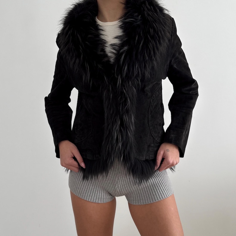 May include: A black jacket featuring a faux fur collar and trim. The jacket has a suede-like texture and a tailored fit. The collar and trim are a dark, textured faux fur. The jacket is worn over a white top and grey shorts.