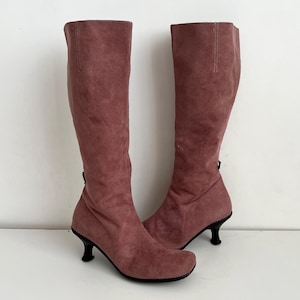 May include: A pair of knee-high boots in a dusty rose color. The boots have a soft, suede-like texture and feature a small, curved heel. A zipper is visible on the back of each boot. The boots are designed for fashion and style.