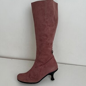 May include: A knee-high, dusty rose-colored boot with a rounded toe and a small, black, curved heel. The boot is made of a suede-like material and features a decorative stitch detail along the back of the shaft.
