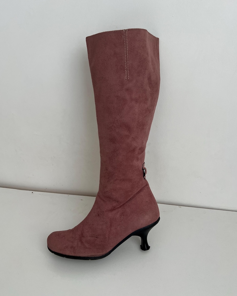 May include: A knee-high, dusty rose-colored boot with a rounded toe and a small, black, curved heel. The boot is made of a suede-like material and features a decorative stitch detail along the back of the shaft.
