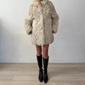 Pu&ograve; includere: Un cappotto di pelliccia vintage, lungo fino al ginocchio, nei toni del crema e del grigio. Il cappotto presenta un motivo a spina di pesce e un ampio colletto. La persona indossa stivali neri al ginocchio.