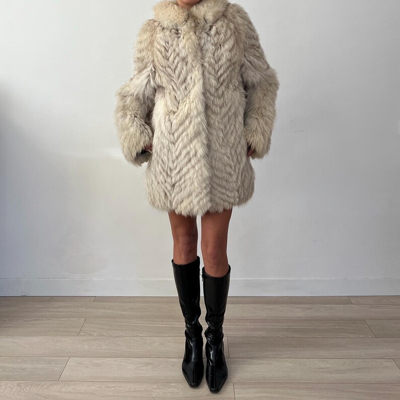 Pu&ograve; includere: Un cappotto di pelliccia vintage, lungo fino al ginocchio, nei toni del crema e del grigio. Il cappotto presenta un motivo a spina di pesce e un ampio colletto. La persona indossa stivali neri al ginocchio.