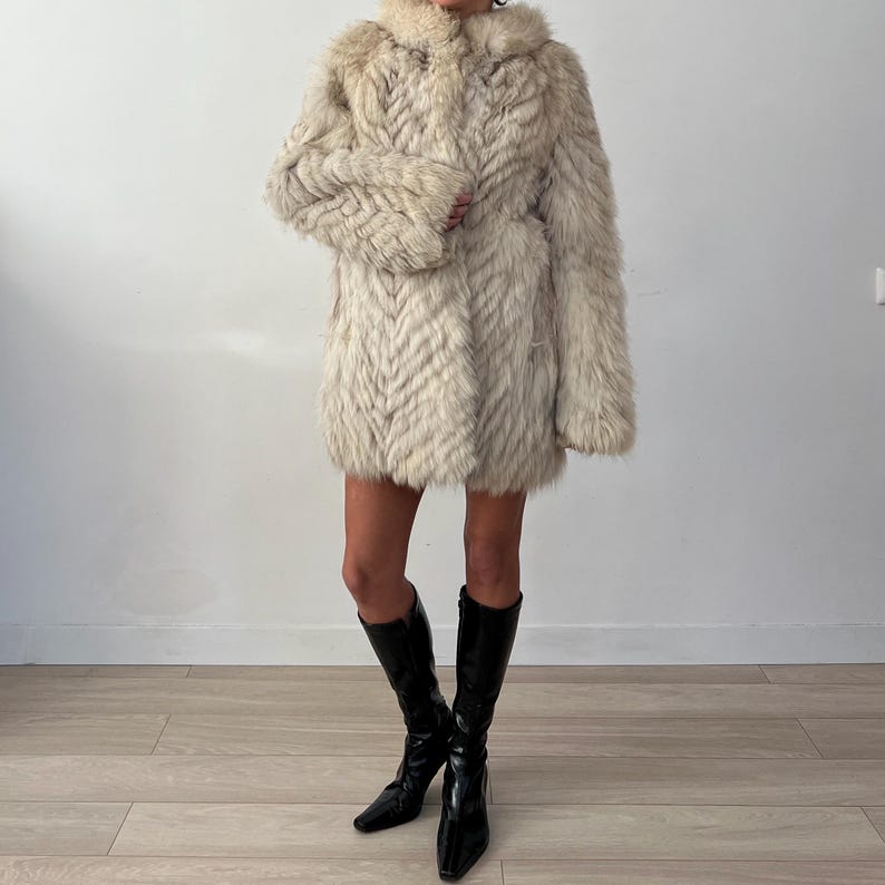 Pu&ograve; includere: Un cappotto di pelliccia color crema, lungo fino al ginocchio, con un motivo a spina di pesce. Il cappotto ha un colletto e maniche lunghe. La persona indossa stivali neri al ginocchio.