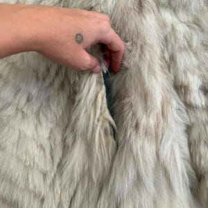 Pu&ograve; includere: Primo piano di un cappotto di pelliccia colorata. La pelliccia &egrave; un mix di bianco e marrone chiaro, con fili lunghi e morbidi. Il cappotto sembra essere vintage o usato, con qualche segno di usura visibile. La consistenza della pelliccia &egrave; evidente, creando una sensazione lussuosa.