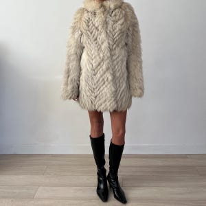 Pu&ograve; includere: Un cappotto di pelliccia colorata, lungo fino al ginocchio, con un motivo strutturato a forma di chevron. Il cappotto ha un ampio colletto e maniche lunghe. La persona indossa stivali neri al ginocchio.