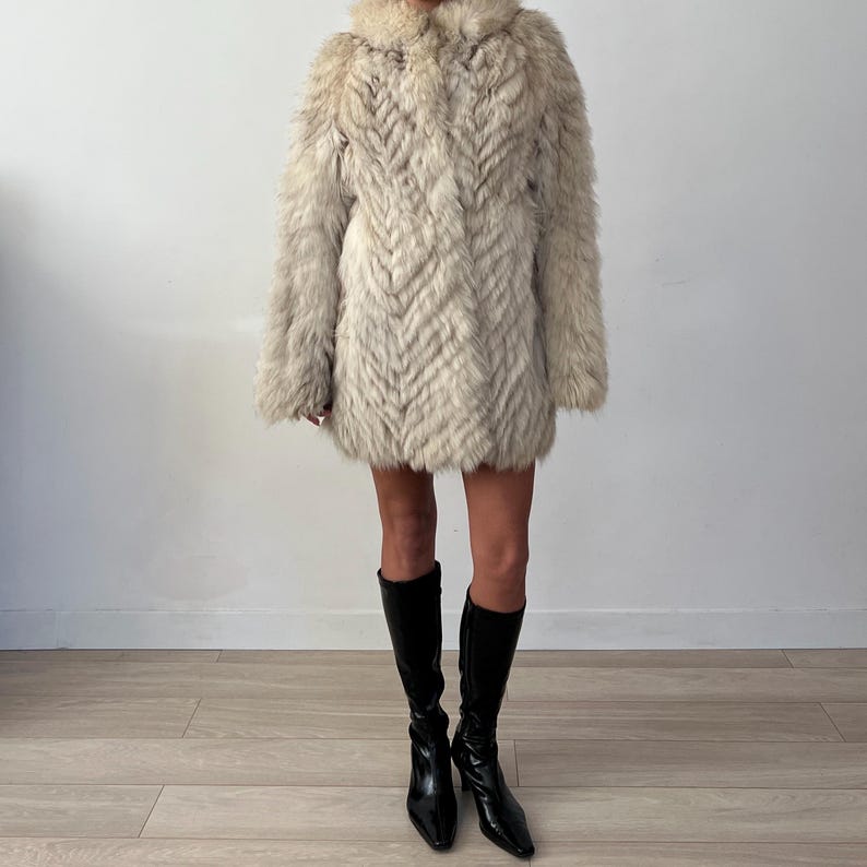 Pu&ograve; includere: Un cappotto di pelliccia colorata, lungo fino al ginocchio, con un motivo strutturato a forma di chevron. Il cappotto ha un ampio colletto e maniche lunghe. La persona indossa stivali neri al ginocchio.