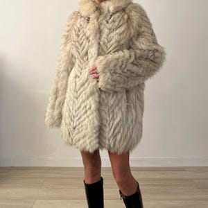Pu&ograve; includere: Un lungo cappotto di pelliccia color crema con un motivo a spina di pesce. Il cappotto ha un ampio colletto e maniche lunghe. La persona indossa stivali neri. Il cappotto sembra essere realizzato in un materiale morbido e lussuoso.
