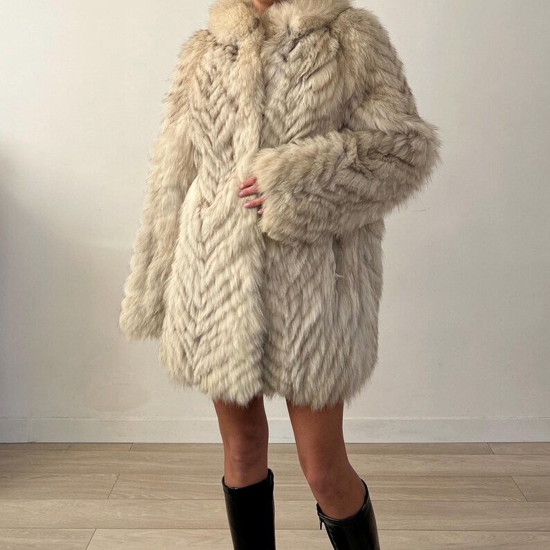 Pu&ograve; includere: Un lungo cappotto di pelliccia color crema con un motivo a spina di pesce. Il cappotto ha un ampio colletto e maniche lunghe. La persona indossa stivali neri. Il cappotto sembra essere realizzato in un materiale morbido e lussuoso.