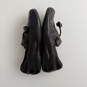 Puede incluir: Un par de zapatos de cuero negro con punta redondeada y cierre de correa. Los zapatos tienen una suela texturizada y un detalle de cord&oacute;n en el tal&oacute;n. Los zapatos se muestran sobre un fondo blanco.