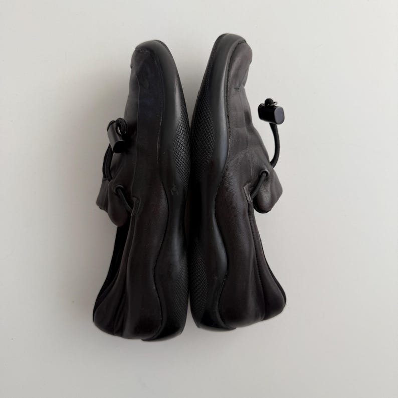 Puede incluir: Un par de zapatos de cuero negro con punta redondeada y cierre de correa. Los zapatos tienen una suela texturizada y un detalle de cord&oacute;n en el tal&oacute;n. Los zapatos se muestran sobre un fondo blanco.