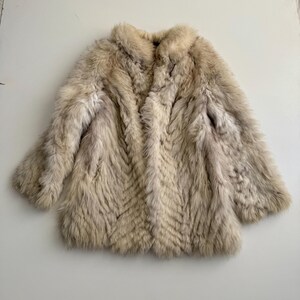 Pu&ograve; includere: Un cappotto di pelliccia colorata, con la parte anteriore aperta e un motivo strutturato a forma di chevron. Il cappotto ha maniche lunghe e un colletto. La pelliccia sembra essere un mix di marrone chiaro e crema, creando un aspetto morbido e lussuoso. Il cappotto &egrave; appoggiato su una superficie bianca.
