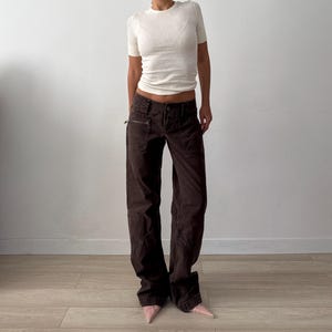 May include: A white short-sleeved crew neck t-shirt paired with dark brown wide-leg trousers. The trousers feature a low-rise waist, multiple pockets including a zippered side pocket, and visible seam detailing along the legs. Light pink pointed shoes.