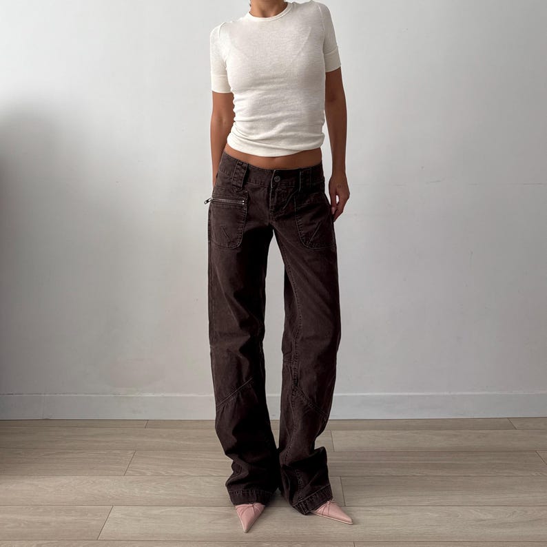 May include: A white short-sleeved crew neck t-shirt paired with dark brown wide-leg trousers. The trousers feature a low-rise waist, multiple pockets including a zippered side pocket, and visible seam detailing along the legs. Light pink pointed shoes.