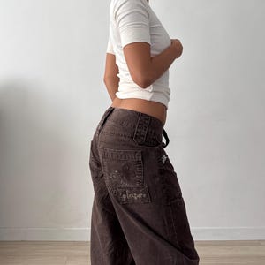 May include: A white short-sleeved ribbed t-shirt paired with dark brown wide-leg pants. The pants feature a low-rise waist, belt loops, and a rectangular patch on the back pocket with the word pleasure embroidered in a cursive script font.