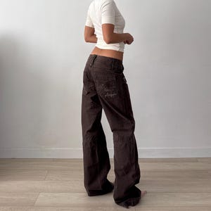 May include: A pair of dark brown low-rise cargo pants featuring a wide-leg silhouette, multiple pockets, and a visible embroidered graphic on the back pocket with the text "pleasures" stitched in a matching dark thread color.