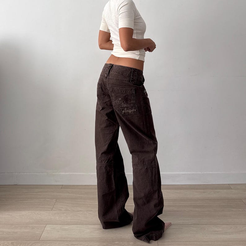 May include: A pair of dark brown low-rise cargo pants featuring a wide-leg silhouette, multiple pockets, and a visible embroidered graphic on the back pocket with the text "pleasures" stitched in a matching dark thread color.