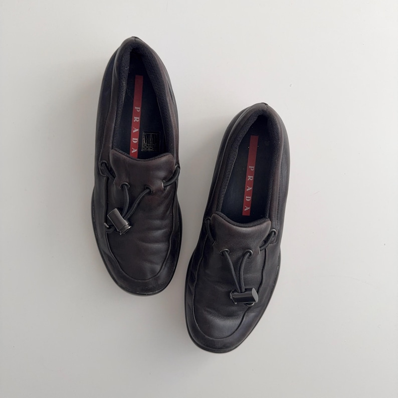 Puede incluir: Un par de mocasines de cuero marr&oacute;n oscuro con un detalle de borla. Los zapatos tienen una puntera redondeada y un dise&ntilde;o sin cordones. El interior del zapato tiene una etiqueta roja con la marca "PRADA" en blanco.