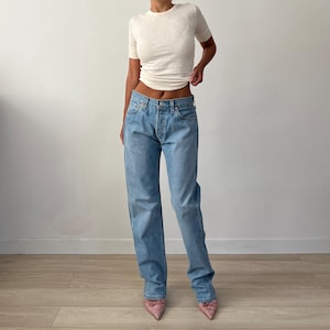 Puede incluir: Vaqueros de mezclilla azul claro con bragueta de botones, combinados con una camiseta de manga corta de color blanco roto. El conjunto se completa con tacones rosas de punta. La modelo está de pie frente a una pared blanca.